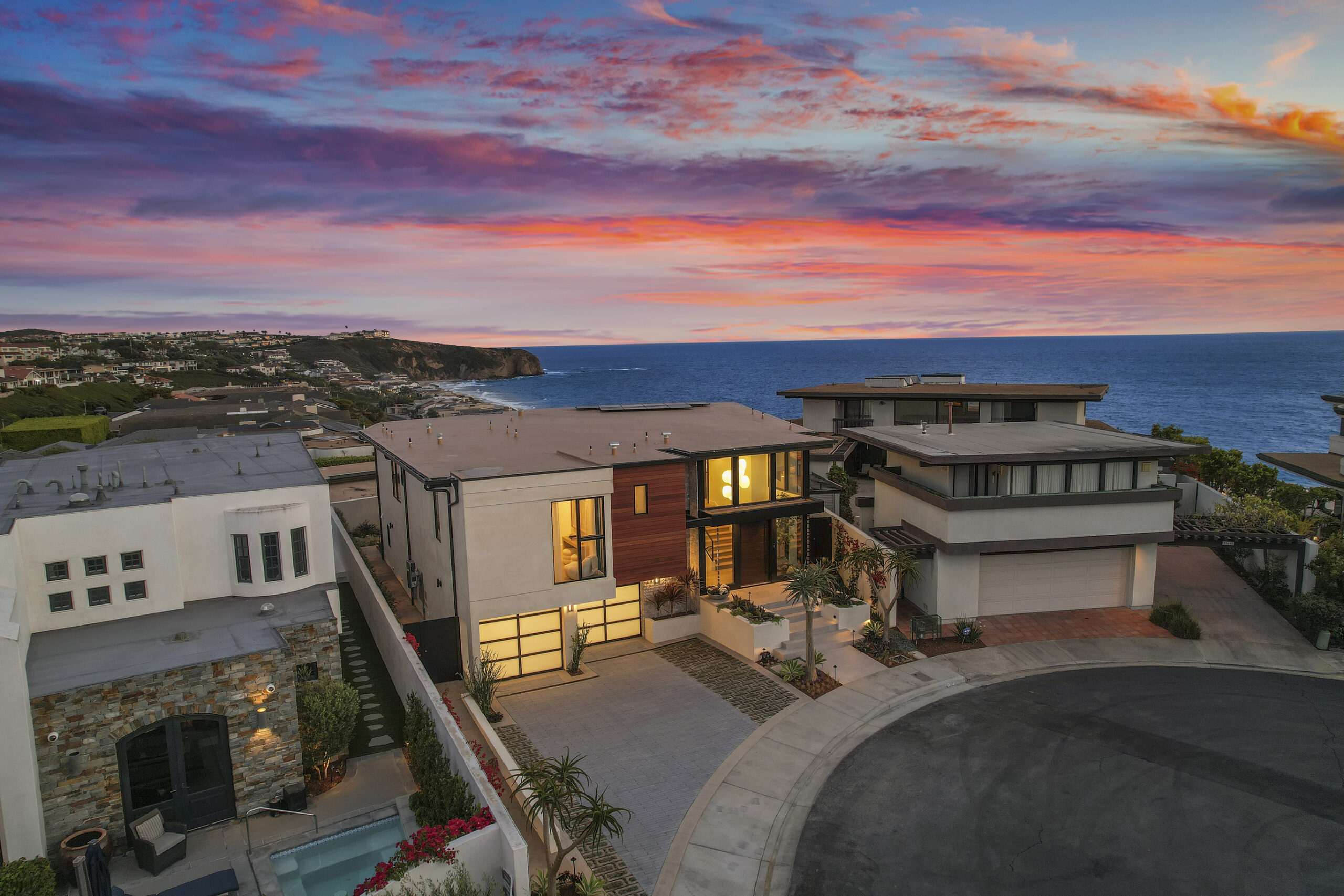 The striking aerial view of this contemporary two-story home blends smooth stucco, natural wood cladding, and textured stone, creating a warm yet modern aesthetic with strong curb appeal.  Sleek glass garage doors with minimalist aluminum framing complement the home's modern lines, adding architectural interest. Stunning sunset ocean view completes the scene.