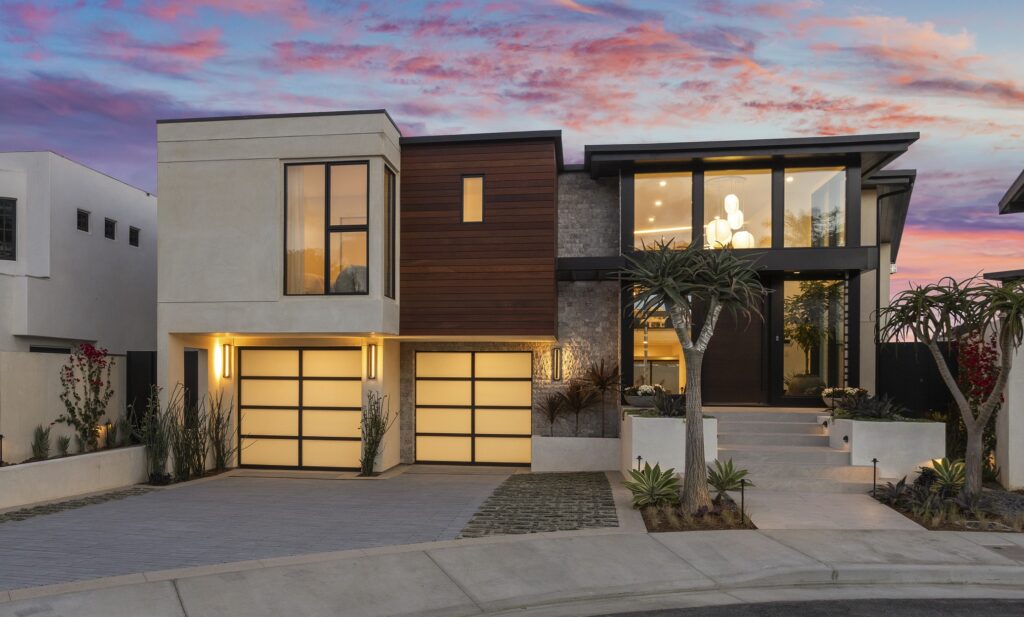 The striking two-story home blends smooth stucco, natural wood cladding, and textured stone, creating a warm yet modern aesthetic with strong curb appeal.  Sleek glass garage doors with minimalist aluminum framing complement the home's modern lines. Shown at twilight.