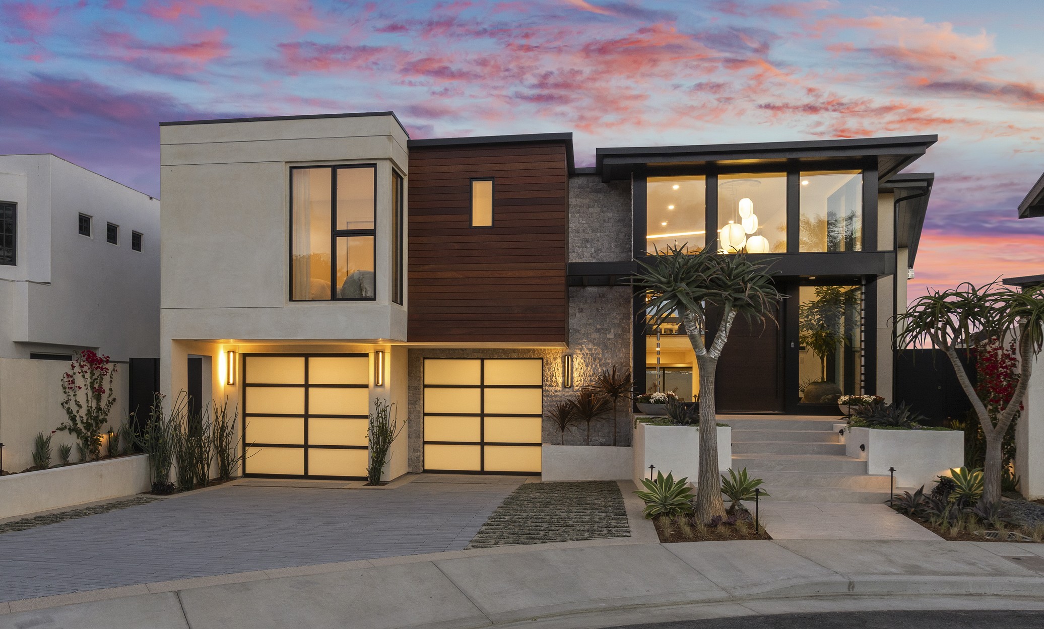 The striking two-story home blends smooth stucco, natural wood cladding, and textured stone, creating a warm yet modern aesthetic with strong curb appeal.  Sleek glass garage doors with minimalist aluminum framing complement the home's modern lines. Shown at twilight.
